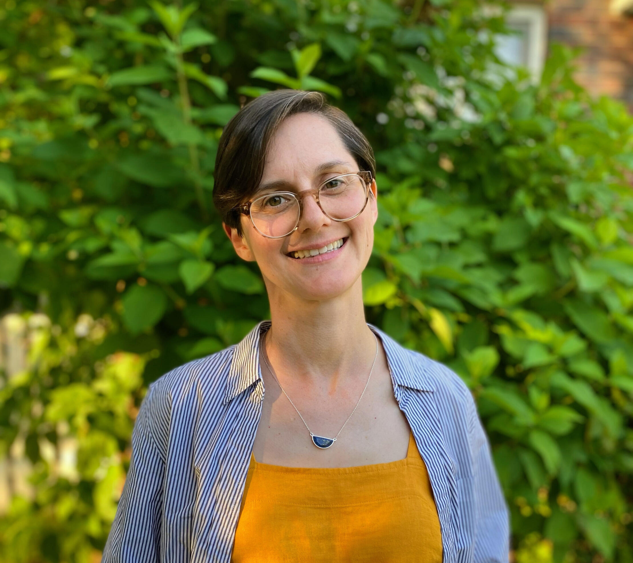 Portrait of a white, non-binary person with short brown and gray hair, standing in front of a green hydrangea bush.